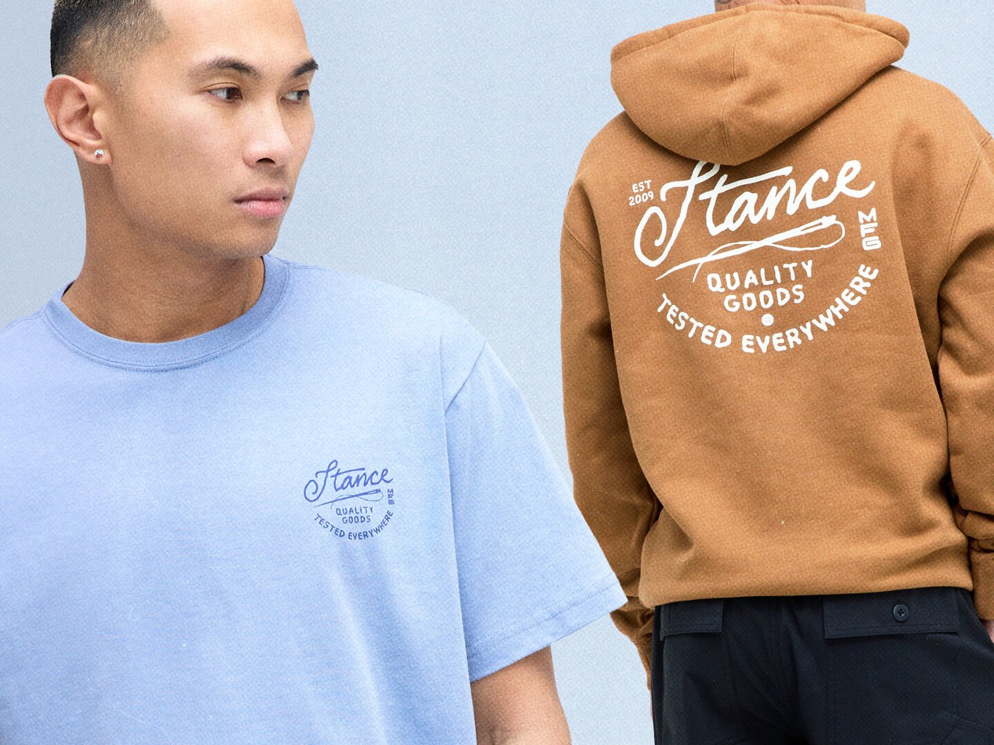 Model wearing a blue t-shirt standing in front of another model in a saddle colored hoodie